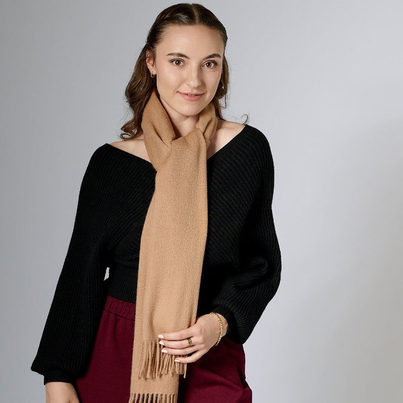 A woman wearing a beige camel colored scarf with fringes, paired with a black top and red pants.