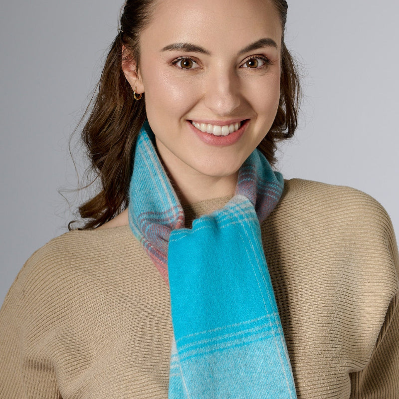 A woman smiling, wearing a blue, red, and Gray plaid scarf with a beige jacket and a light-colored top.