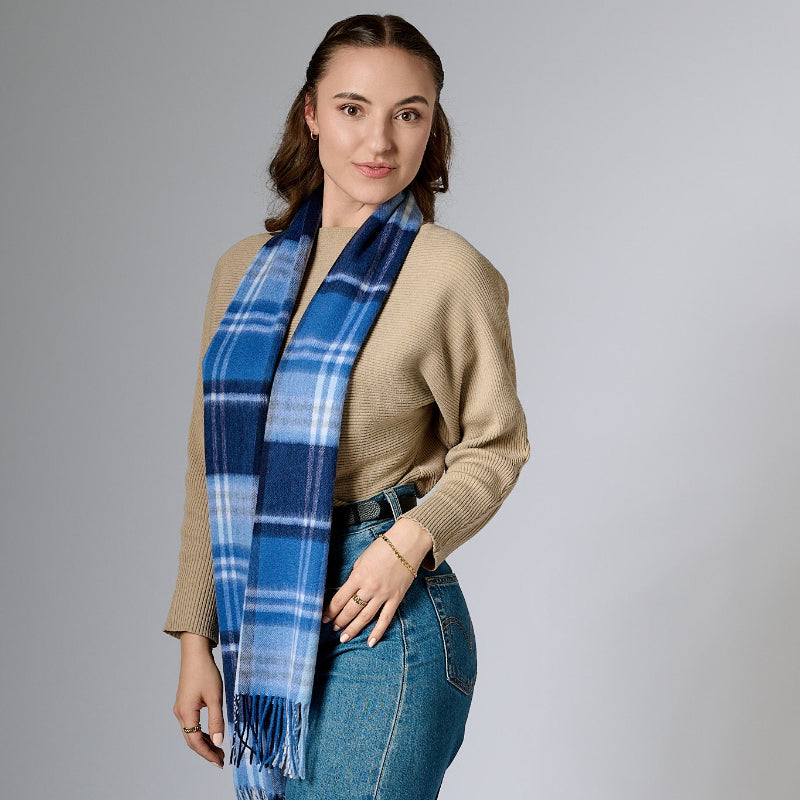 A woman wearing a beige knit top and blue jeans, accessorized with a blue checkered-patterned merino wool scarf and a brown belt.