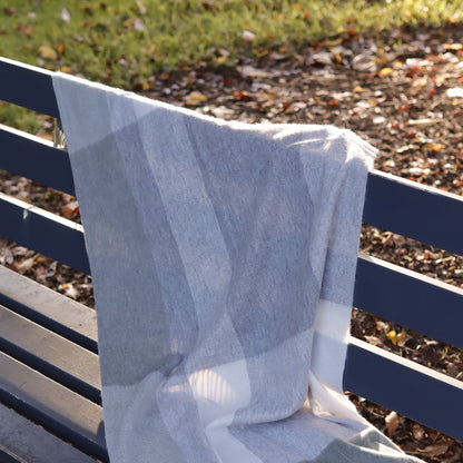 A light gray and white plaid throw blanket draped over a dark blue wooden park bench, with a blurred outdoor background of grass and fallen leaves.