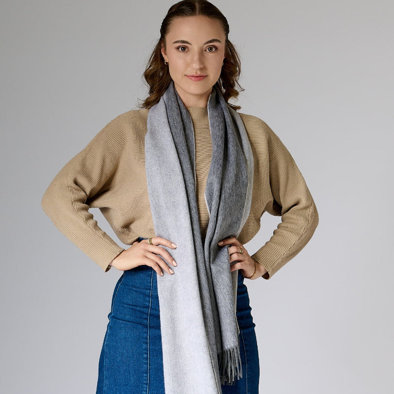 Woman wearing a beige sweater and blue jeans with a gray scarf on a plain background