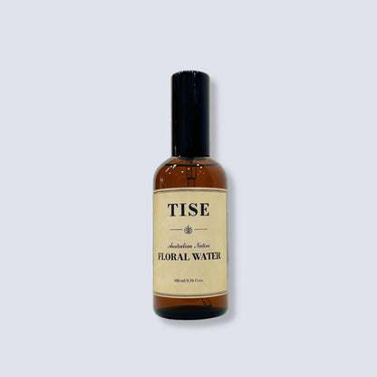 A bottle of TISE Floral Water with an amber glass body, black cap, and cream-colored label, centered on a light gray background.