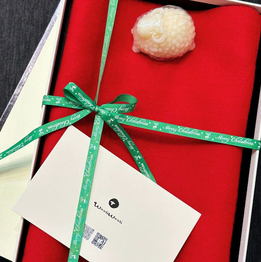 Gift box with green ribbon and a card on a dark surface. A red wool scarf and a handmade goat soap are in this box. 