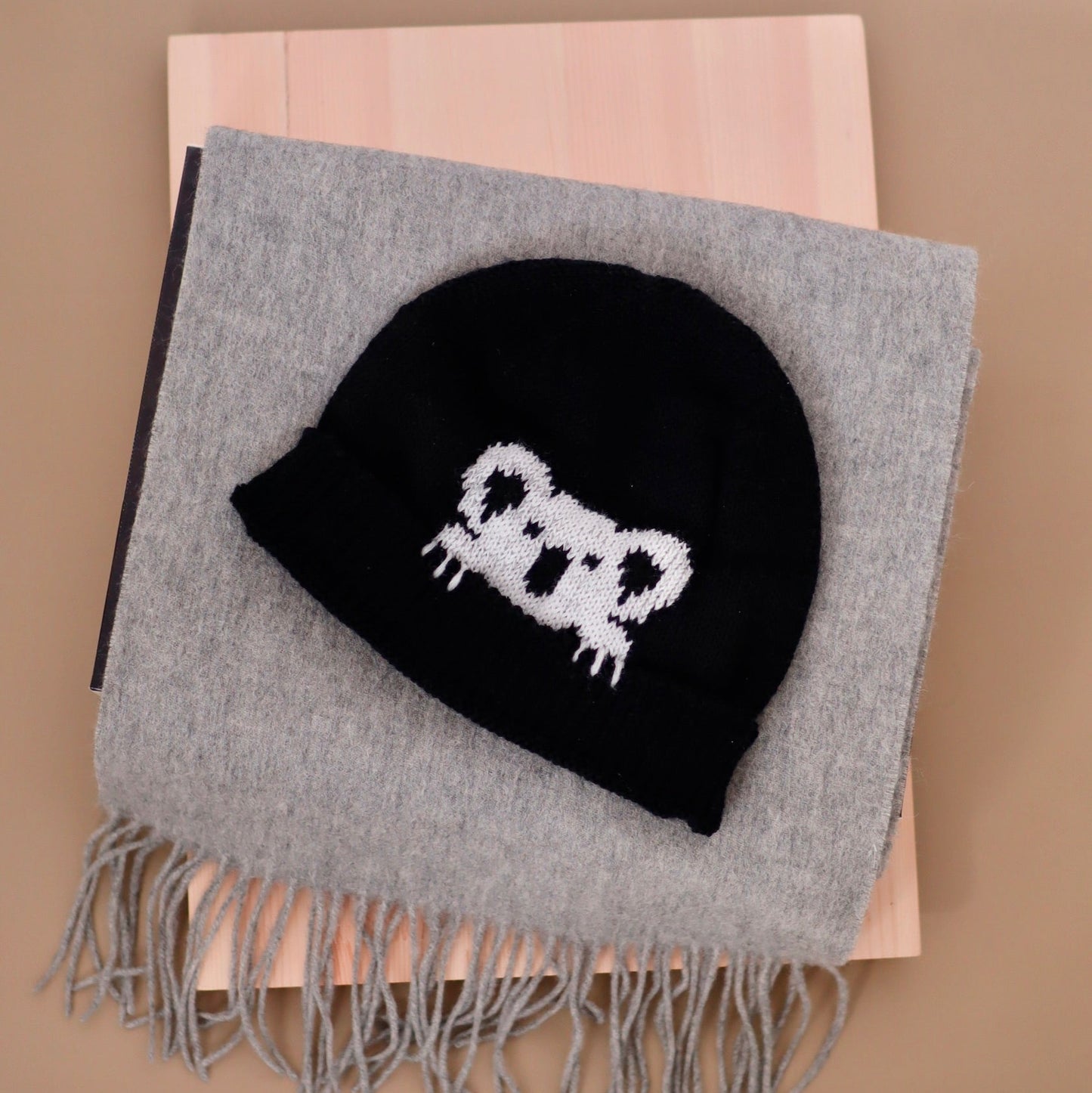A black knit beanie with a white cartoon face motif, displayed on top of a folded gray fringed scarf over a light wood block against a beige background.