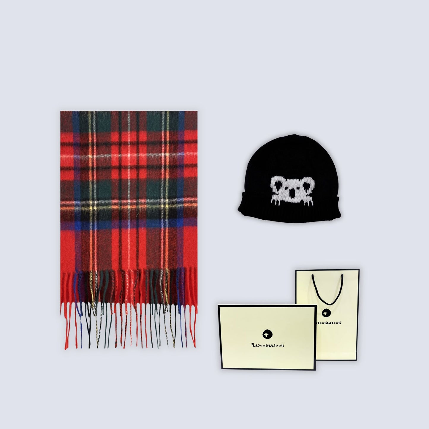 A flat-lay product shot featuring a red and black plaid scarf with fringe, a black beanie with a white koala face logo, and two cream-colored branded paper bags on a light gray background.