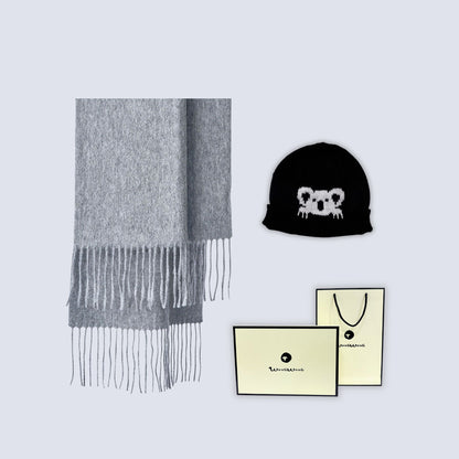 A light gray knit scarf with fringe, a black beanie featuring a white koala face, and cream-colored branded gift packaging on a light gray background.