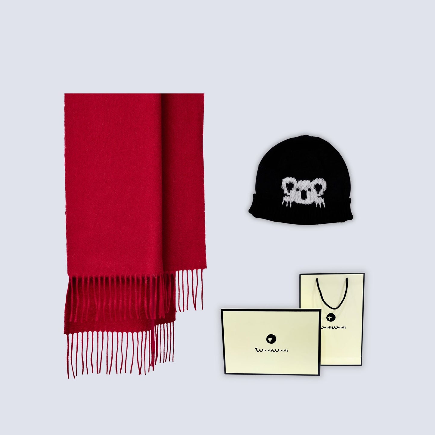 A red knit scarf with fringe, a black beanie featuring a white koala face graphic, and branded cream-colored packaging cards and a shopping bag on a light gray background.