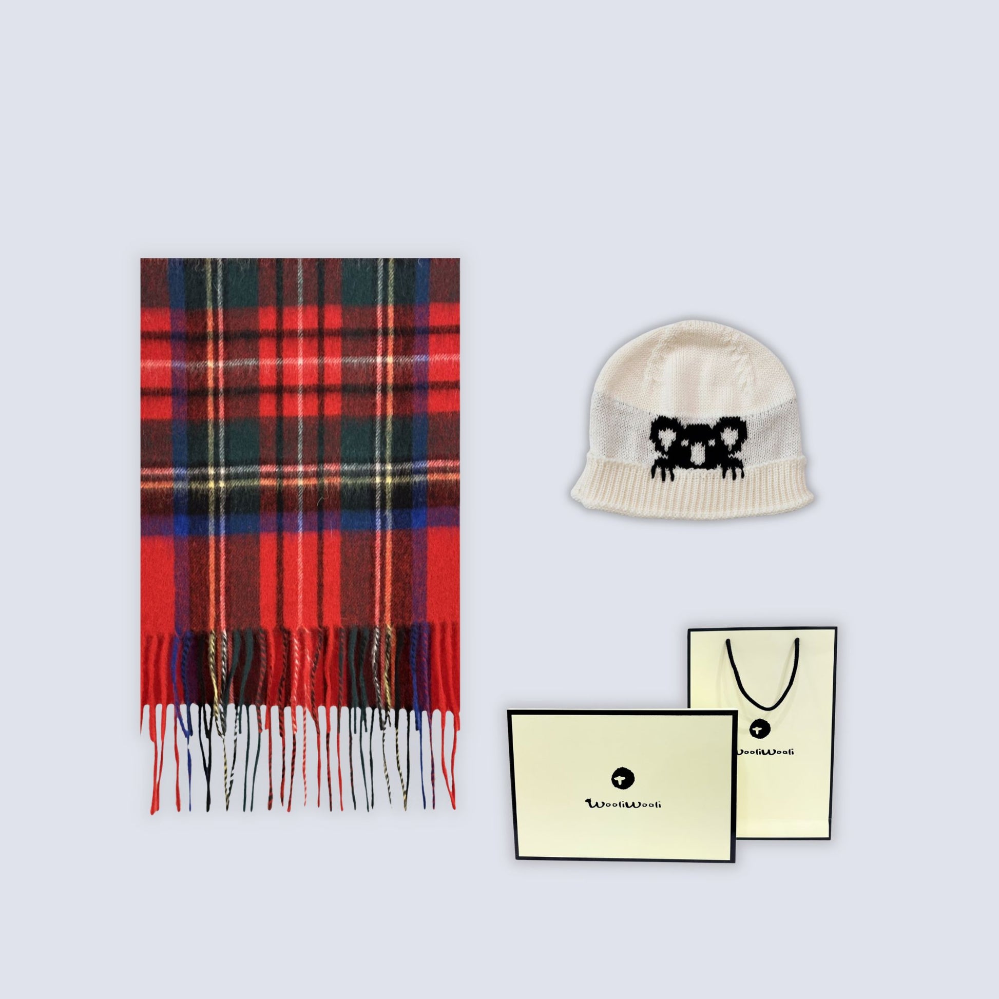 A flat-lay product shot featuring a red and black plaid scarf with fringe, a cream knit beanie with a black embroidered bear face, and two cream-colored branded cards on a light gray background.