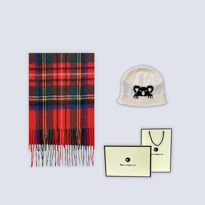 A flat-lay product shot featuring a red and black plaid scarf with fringe, a cream knit beanie with a black embroidered bear face, and two cream-colored branded cards on a light gray background.