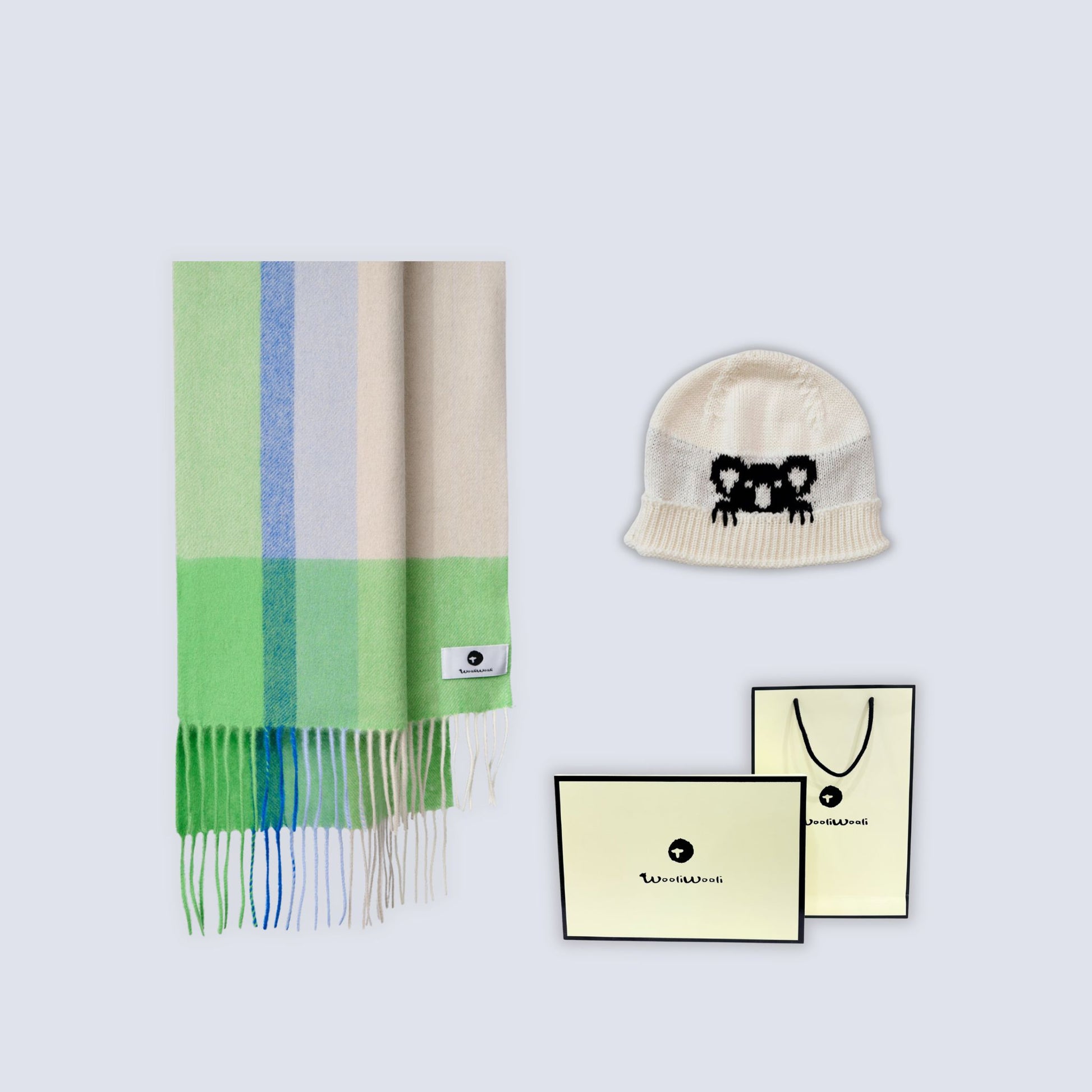 A coordinated knit gift set on a light gray background, including a striped scarf in pale green, blue, cream, and white with fringe, a cream beanie with a black embroidered bear face, and cream branded gift cards and a shopping bag.