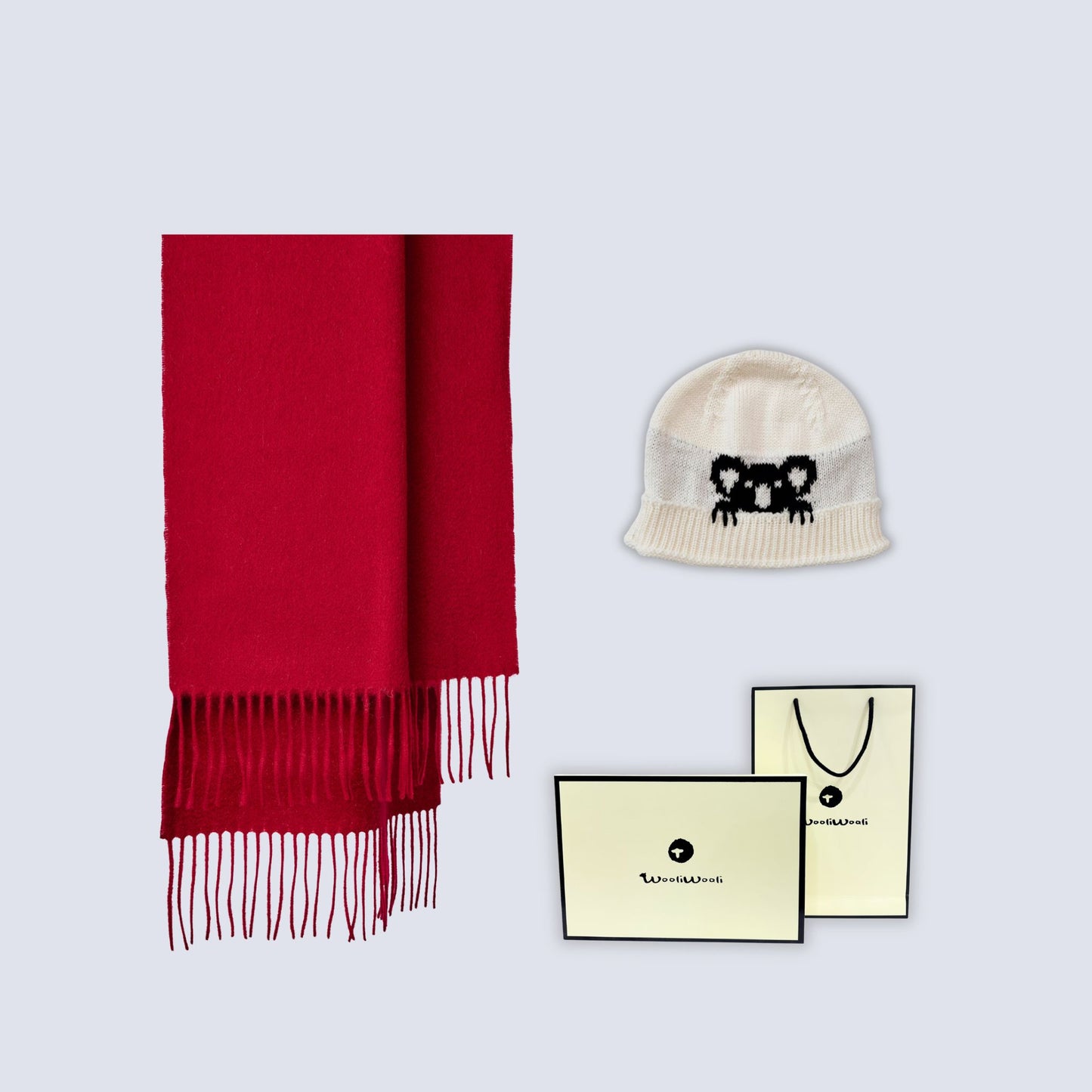 A red knit scarf with fringe, a cream knit beanie featuring a black embroidered bear face, and two cream-colored branded gift bags and cards on a light gray background.