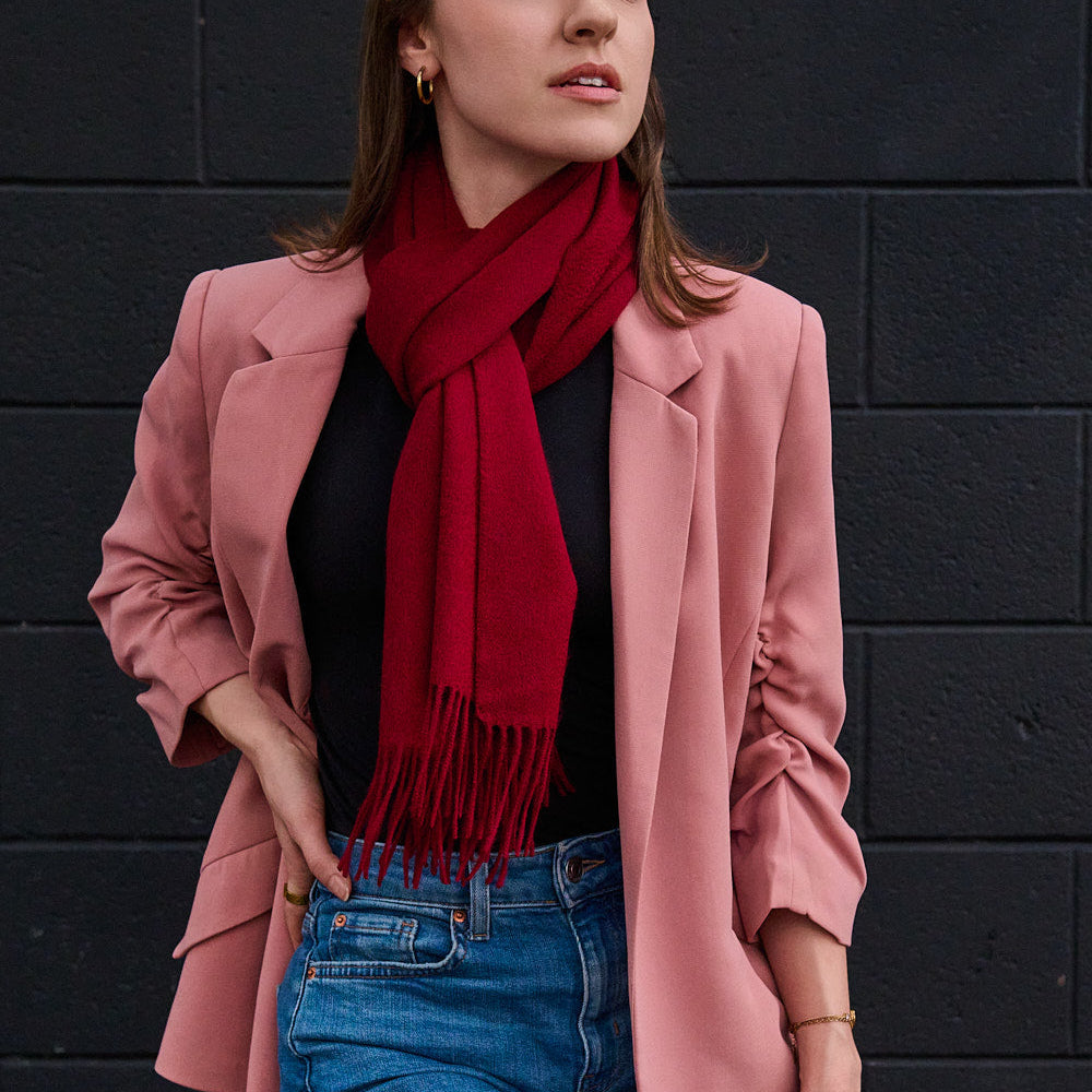A woman wearing a dusty pink blazer, a deep red merino wool scarf with fringe, a black top, and blue jeans, standing against a dark textured brick wall.
